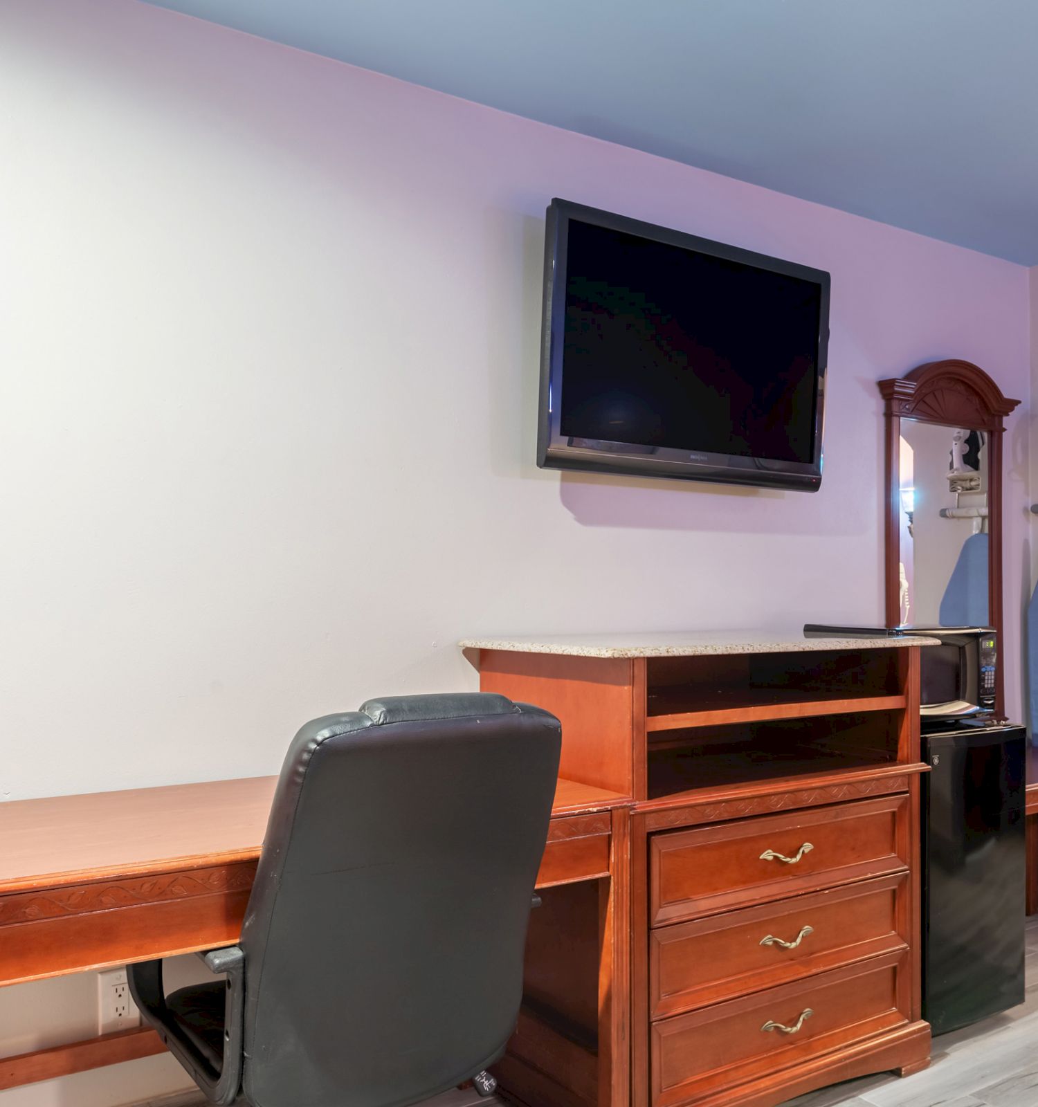 The image shows a hotel room with a desk, chair, TV, dresser, wardrobe, ironing board, and part of a bed visible on the right side.