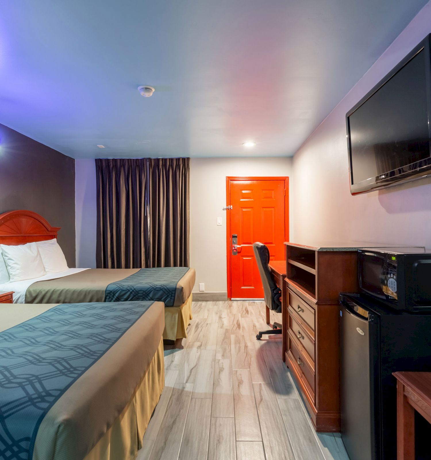 The image shows a hotel room with two beds, a TV, a desk, a microwave, a mini-fridge, and a red door. The room has a modern design and wooden furniture.