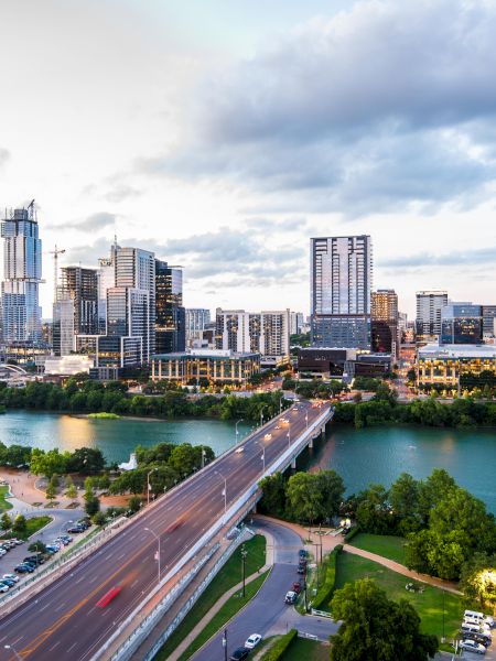 An aerial view of a city skyline featuring tall buildings, a river, a bridge, and surrounding greenery under a partly cloudy sky.