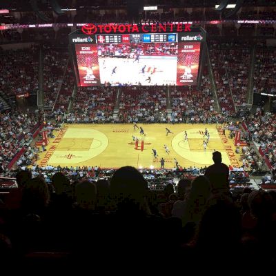 The image shows a basketball game in progress at a stadium with a scoreboard above the court, and the venue is identified as the Toyota Center.