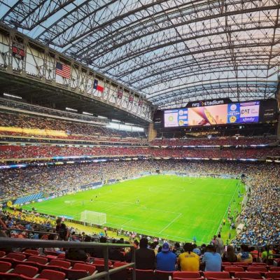 A large stadium with a retractable roof is filled with spectators watching a soccer game on a green field. The atmosphere is lively.