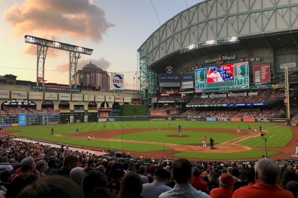 This image shows a baseball game in progress at a stadium with a retractable roof, large crowd, and scoreboard displaying game details.