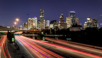 View of a city skyline at night with long exposure light trails from cars on a busy highway in the foreground, creating colorful streaks.