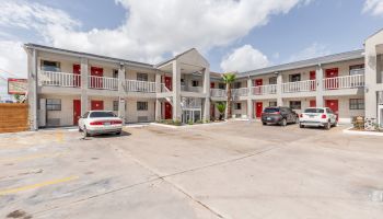 The image shows a two-story motel with red doors, a few cars parked in the lot, and a partially cloudy sky in the background.