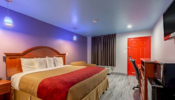 A modern hotel room with a large bed, red door, flat-screen TV, desk, and chair, featuring purple and white walls with hanging light fixtures.