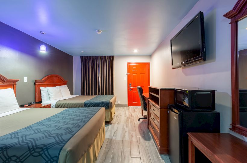 A hotel room with two beds, a red door, TV, desk, chairs, microwave, mini fridge, and a mirror. The room is well-lit with modern decor.